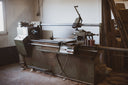 A large industrial metal lathe machine in a workshop or factory setting, with a control panel, carriage, and motor visible against a plain wall.

